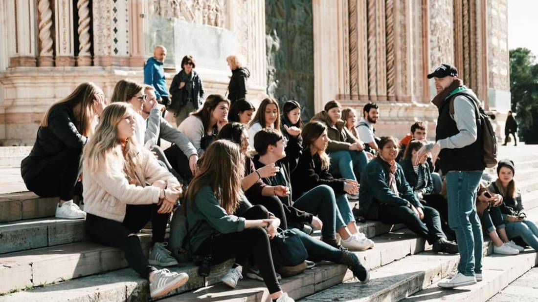 Students sit on the steps outside of the church entrance and listen to man with hat and vest talking.