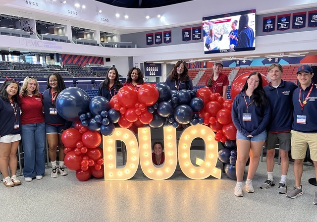 Duke the blue lion poses in front of Duquesne backdrop and balloons.