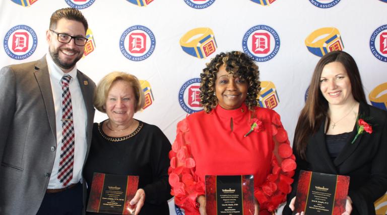 Four people stand holding awards in front of a banner with the Duquesne University alumni association logo 