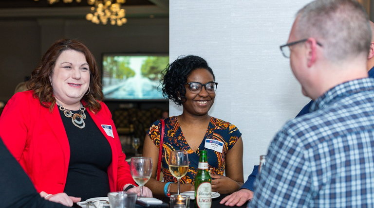 Two people stand talking to other people at a table