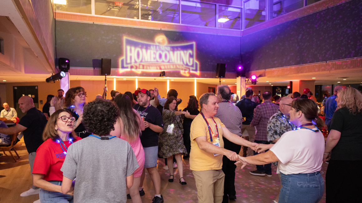 People dancing on a dance floor with the Homecoming & Family Weekend logo projected onto the wall in the background.