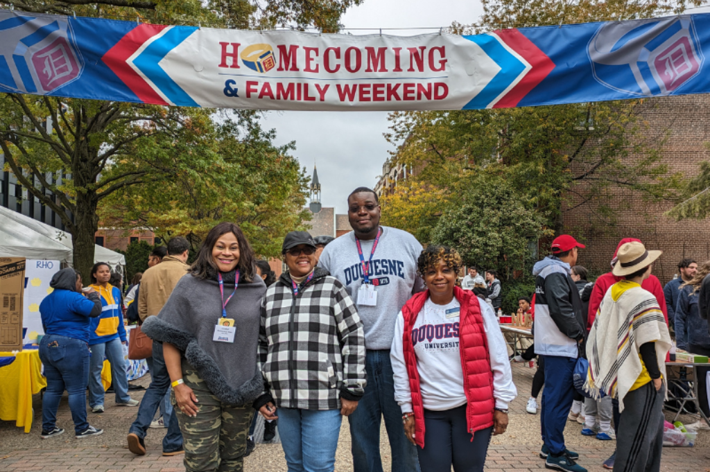 Four people standing together smiling under a banner that says Homecoming & Family Weekend