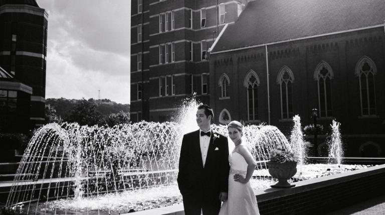 Black and white photo of a bride and groom standing in front of a fountain on Duquesne University's campus with the chapel in the background