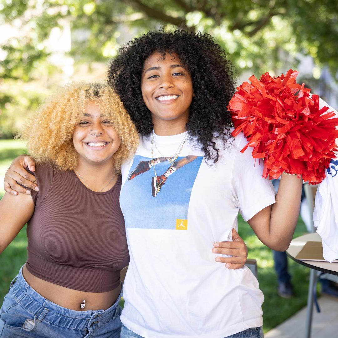 2 Black female students smile at the Unico Pavilion. One of them is holding up a red pom-pom.