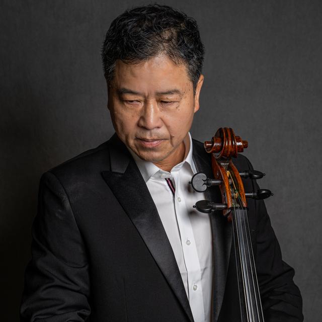 Adam Liu holds a cello and looks away from the camera in front of gray background.
