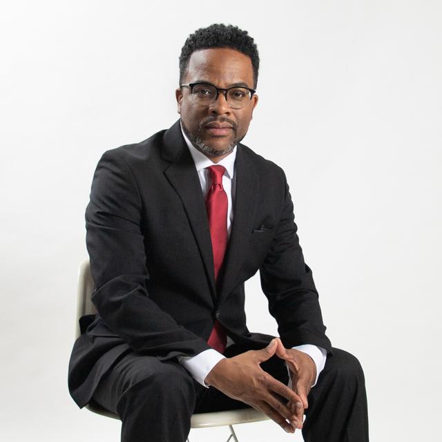 a person posing in front of a white background wearing a black suit with a red tie.