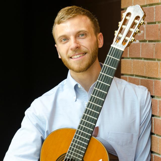 Ben Meyer holds a classical guitar in front of a brick wall.
