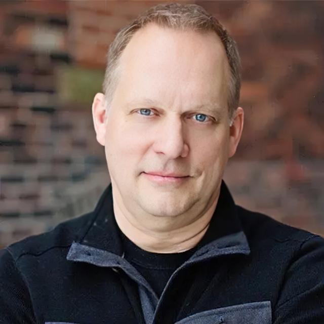 Brent Weber poses for a headshot against a blurry brick wall.