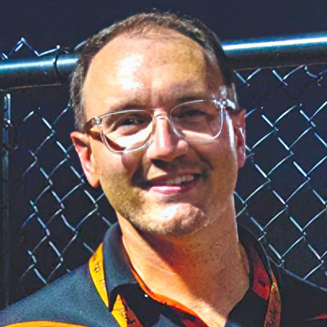 Chad Thompson poses for a headshot outdoors against a chain-link fence.