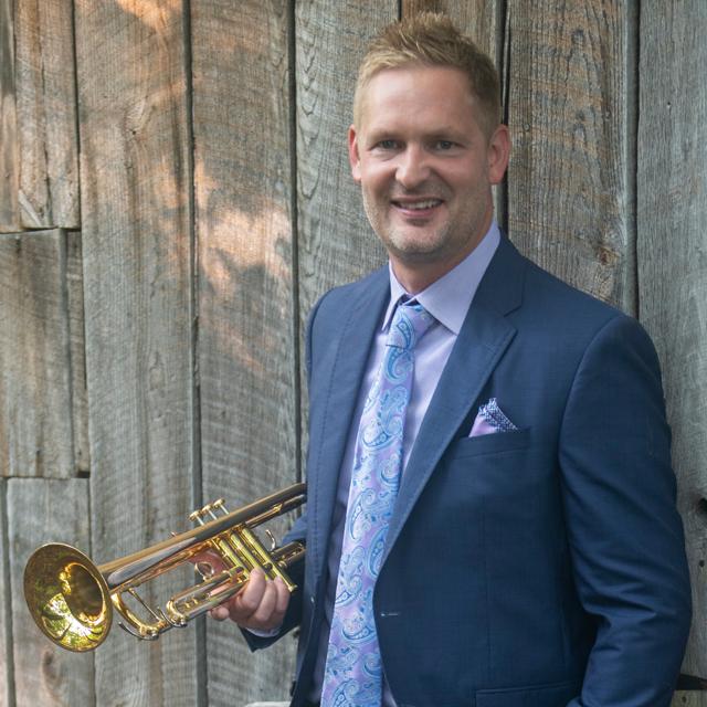 Christopher Wilson poses with trumpet in front of a wooden fence.