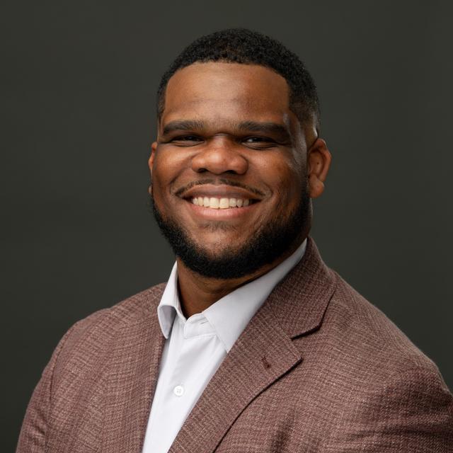 Ethan is smiling, wearing a brown-red blazer and white shirt, against a dark grey solid background.