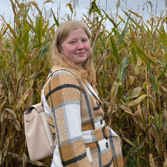Hannah stands in a fieldwork of tall corn in a brown plaid coat.
