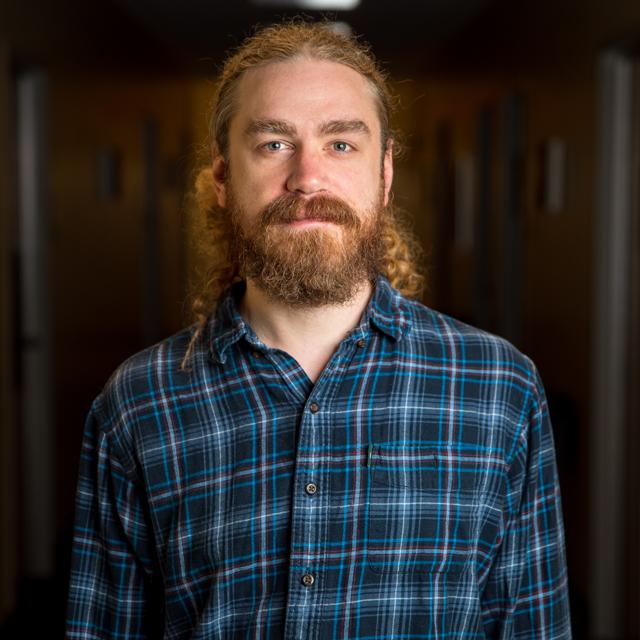 Justin Stag poses for a headshot against a blurry, dark background.