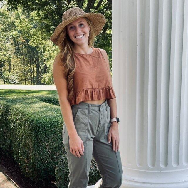 Lauren stand outside smiling in green pants, orange top, and a sun hat.