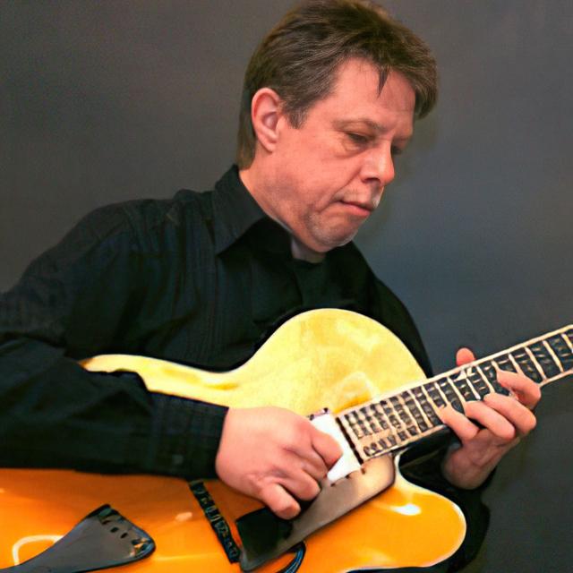 Mark Koch plays a guitar in front of a gray background.