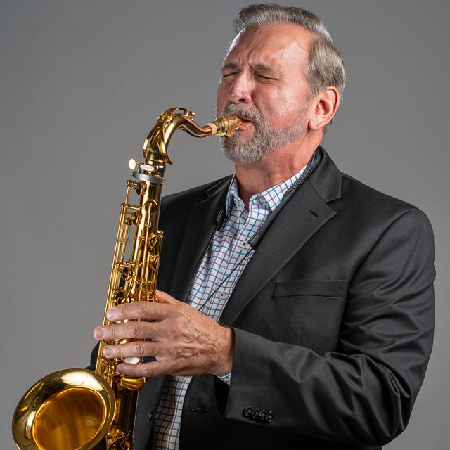 Mike Tomaro plays a saxophone in front of a gray background.