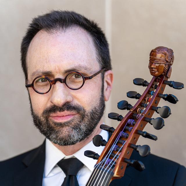 Paul Miller holds a string instrument and poses in front of a beige background.