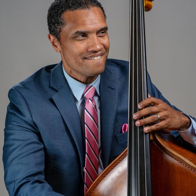 Paul Thompson plays bass in front of a gray backdrop.