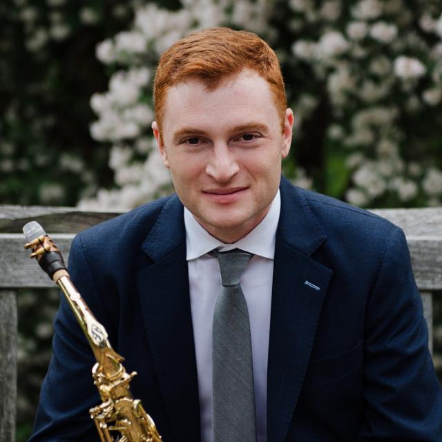 Perry is sitting behind white flowers in a suit, smiling and holding a saxophone.
