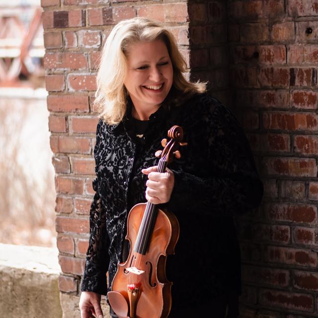Rachel Stegeman holds a violin in front of a brick wall.