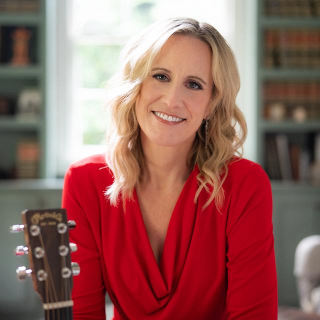 Rachel Whitcomb poses for a photo wearing a red top in a room with bookshelves and an acoustic guitar..