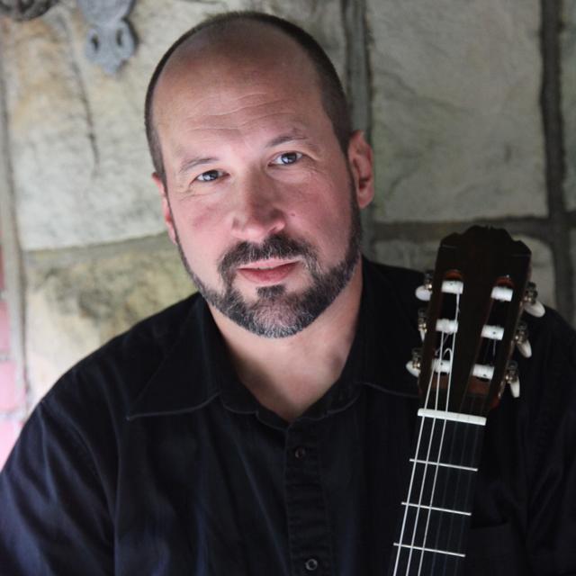 Thomas Kikta holds a classical guitar in front of a brick wall.
