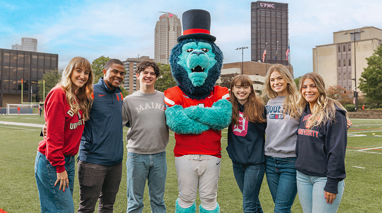 Six Duquesne students standing on Rooney Field with the University's lion mascot.