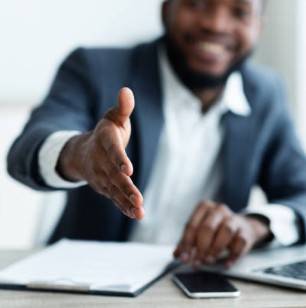 A young black male dressed in professional attire smiles and extends his hand in gesture for a handshake