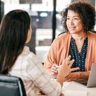 Two females dressed in business casual talk at a table in a professional setting