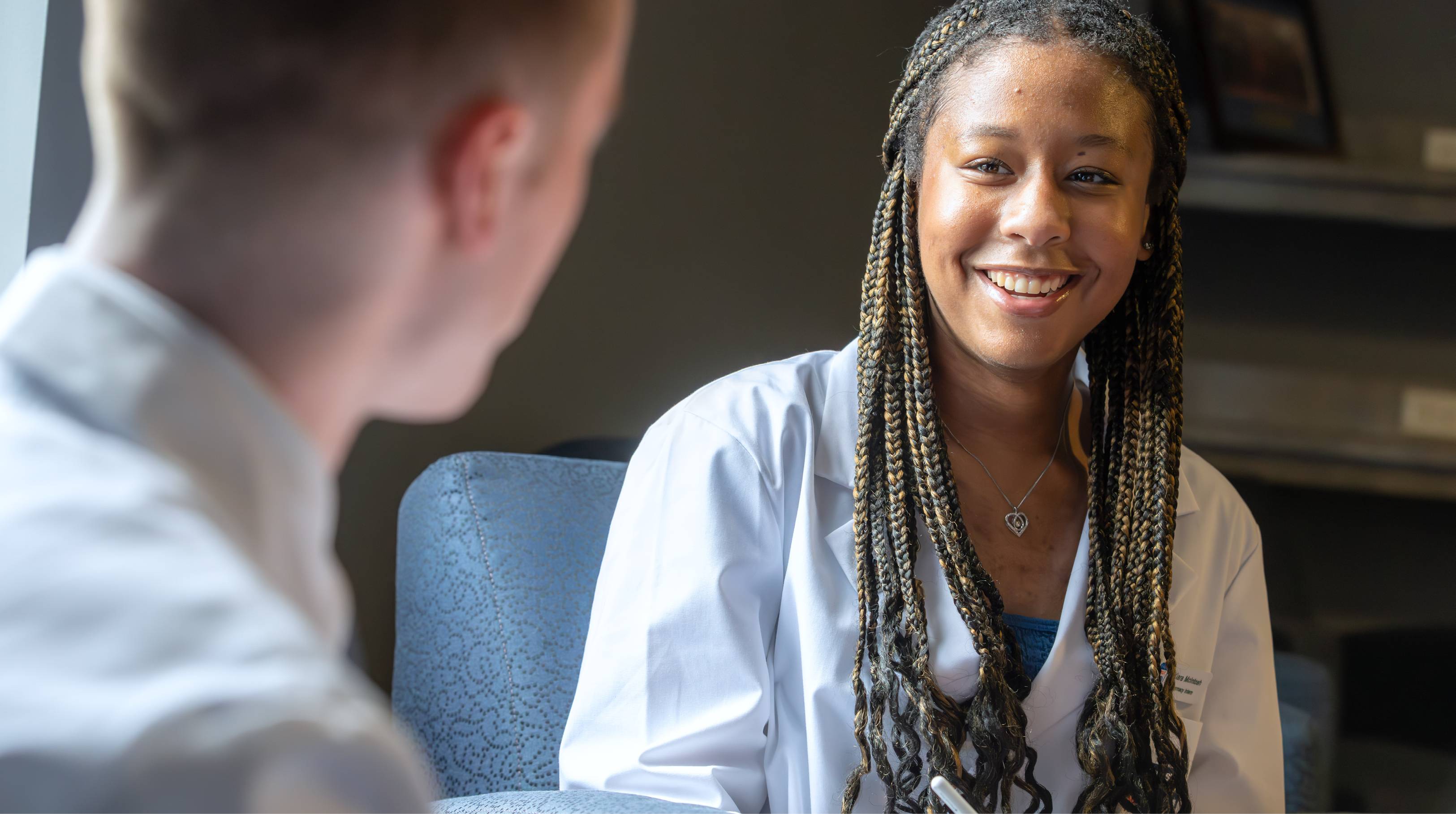 A pharmacy student is smiling and interacting with a pharmacy professional, whose face we cannot see in this image.