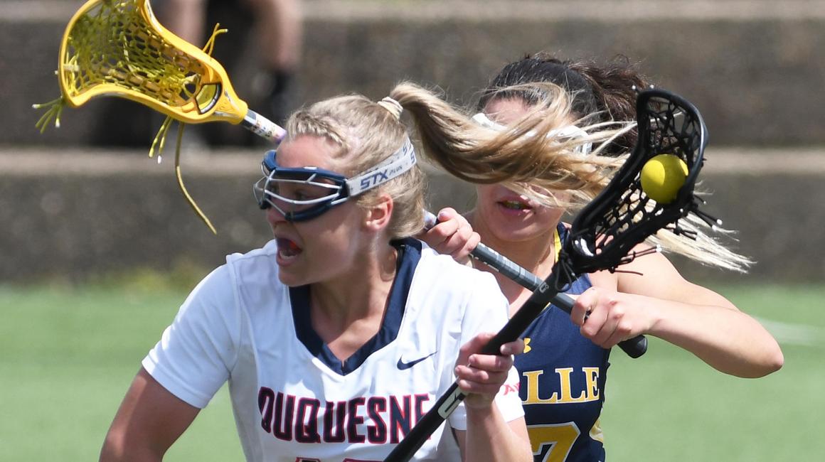 two women's lacrosse players actively playing in a game, Duquesne player in the forefront
