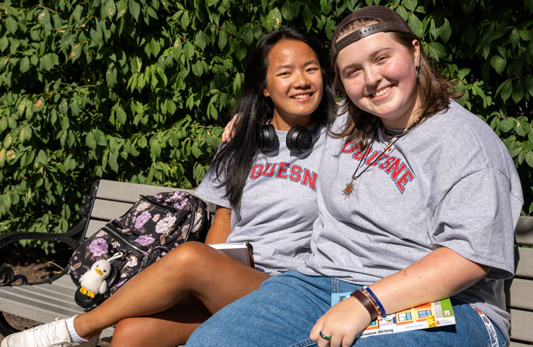 Maia McIntyre and her roommate relax on a bench on campus on a sunny day.