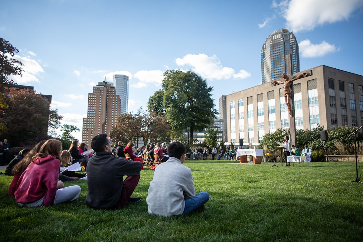 Students sit on the ground outside as the priest celebrates mass