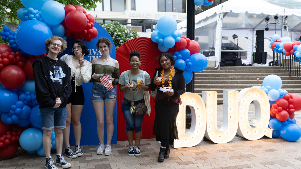 Students smiling on A-Walk surrounded by red and blue balloons and DUQ lettering.