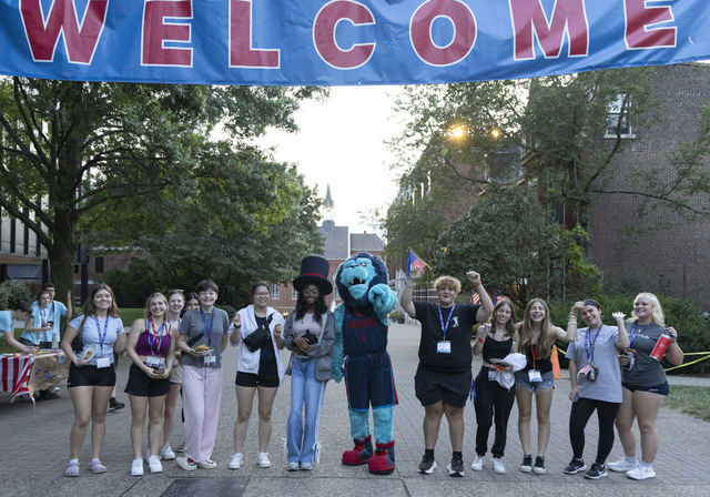 Students and the University mascot stand as a group on campus, beneath the Welcome banner.