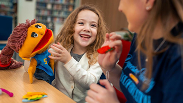 A child sits with a student teacher at a table and plays with a puppet