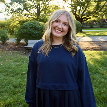 Elle Bowers stands outdoors and smiles for the camera wearing a blue sweater. 
