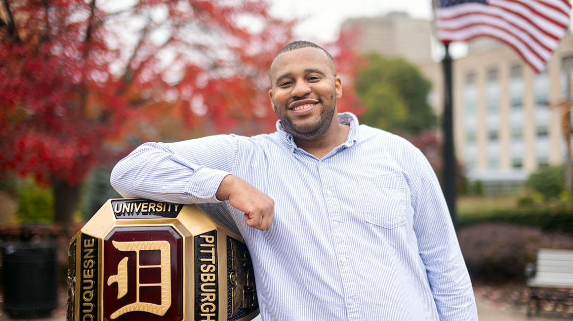 Jeffrey Brown on campus standing next to DU ring with fall tree and American flag in the background.