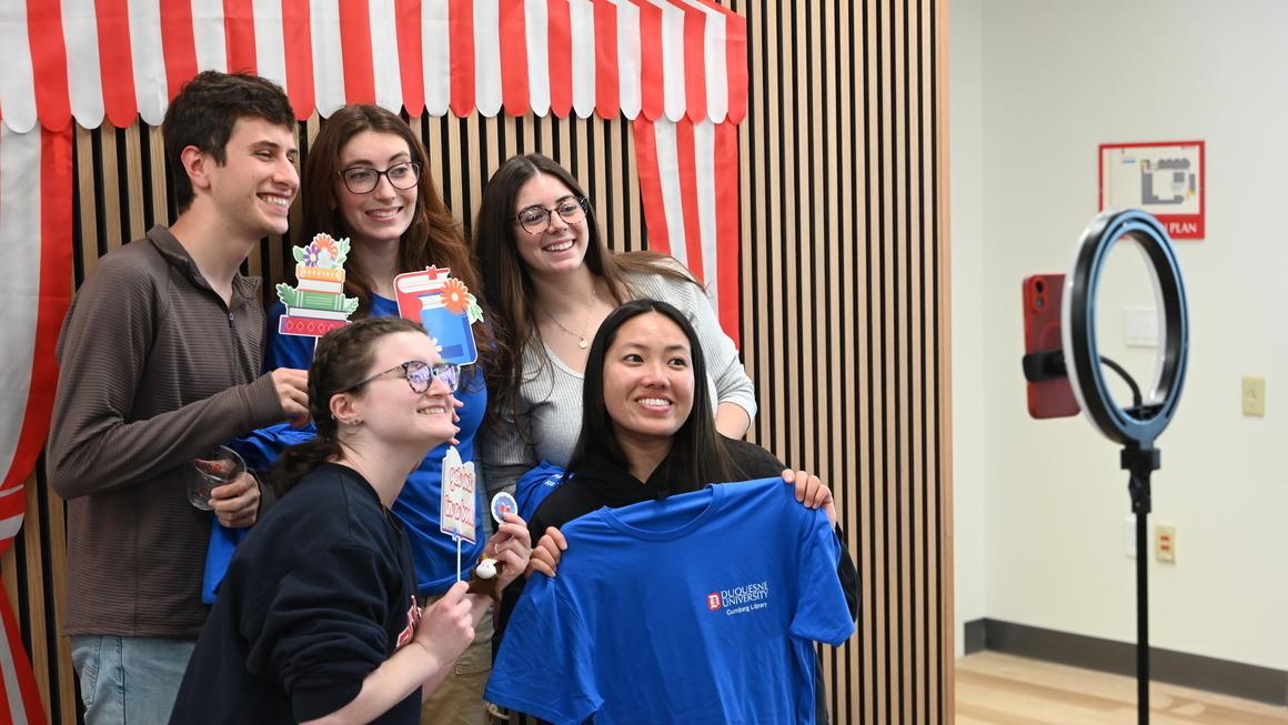 A group of students poses with props and a blue Duquesne t-shirt at the photo booth station. 