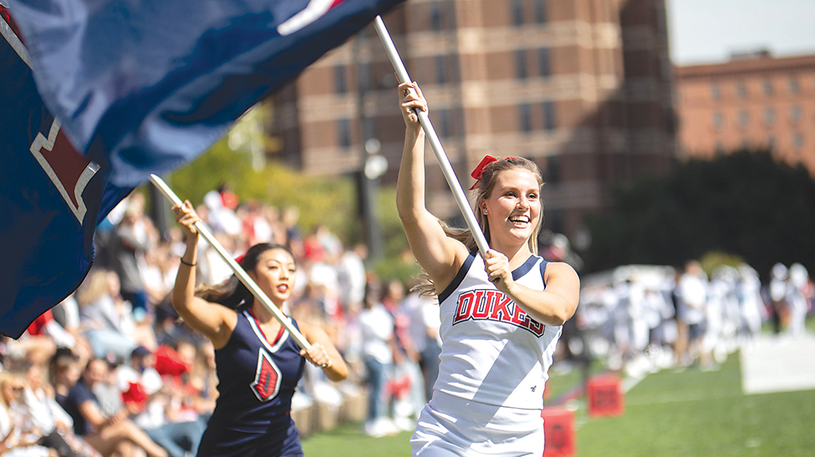 Cheerleaders running on football field with Duquesne flags.