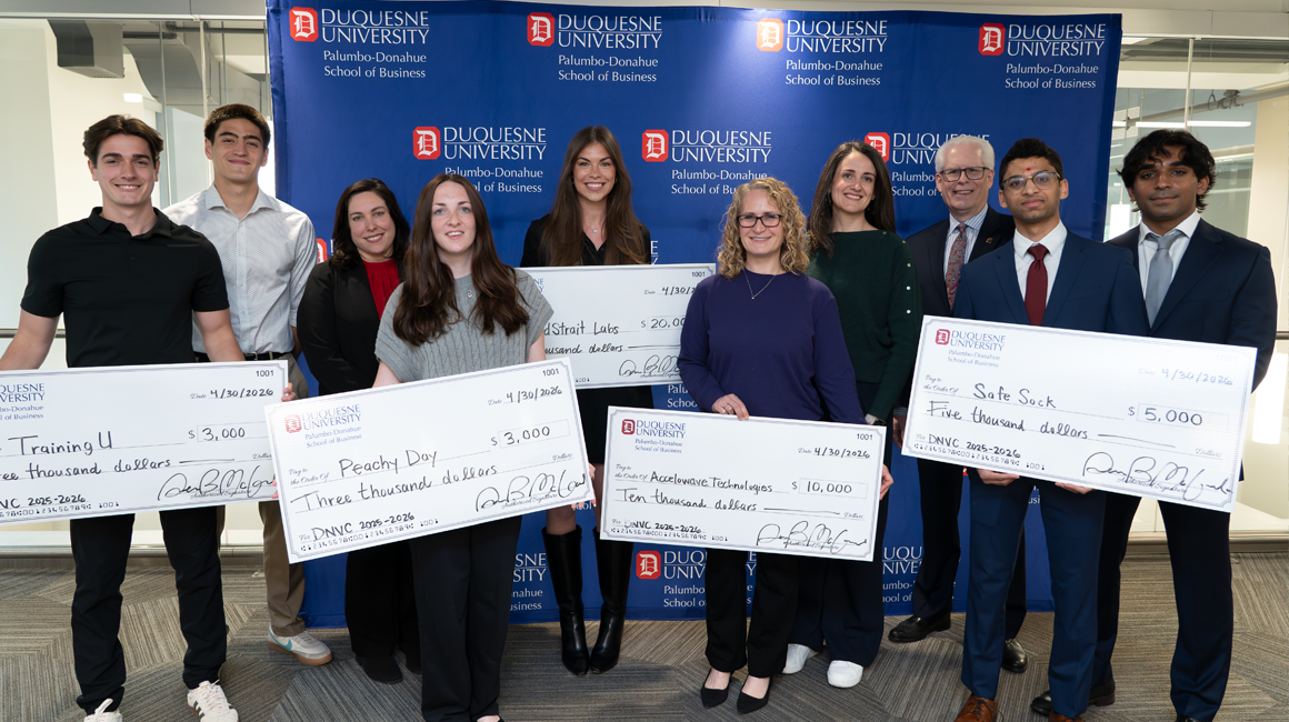 Previous winners of the DNVC pose in front of a Duquesne backdrop holding large prize checks.