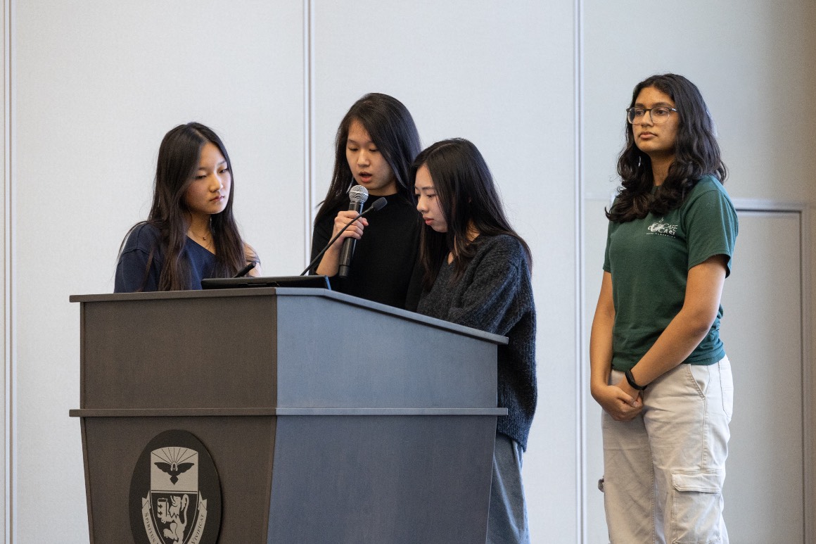 4 students present onstage at lectern at Hacking4Humanity 2026
