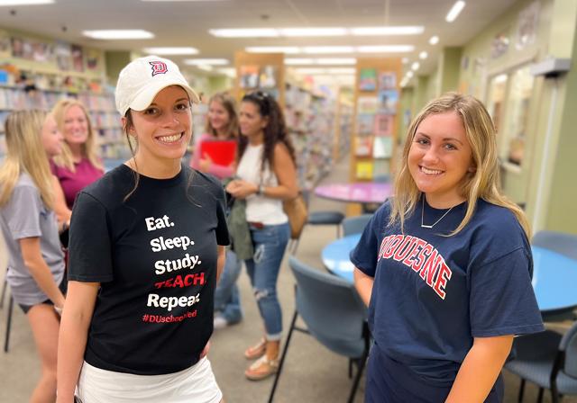 Two School of Education student ambassadors smiling next to each other wearing Duquesne t-shirts and gear with family on admission tour behind them as all are in the library's curriculum center