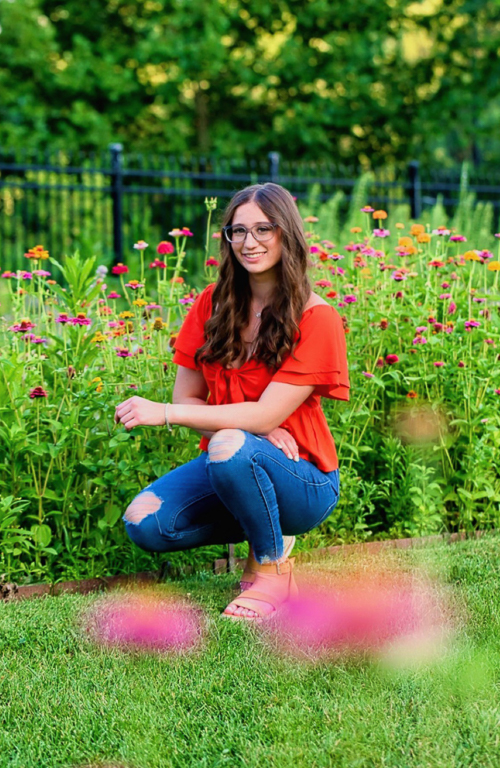 B.S.Ed. Early Childhood Education student Megan Caskey kneeling between wildflowers in field and a fence behind her.
